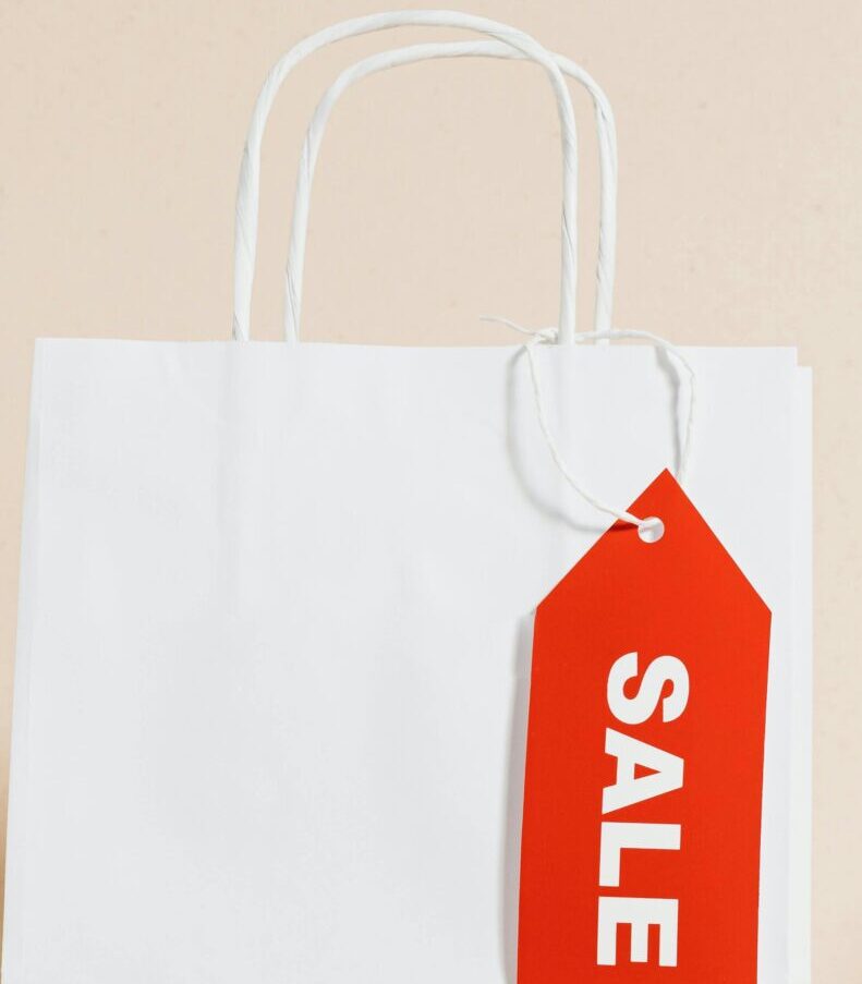 White shopping bag with red sale tag and mini shopping cart on beige background.